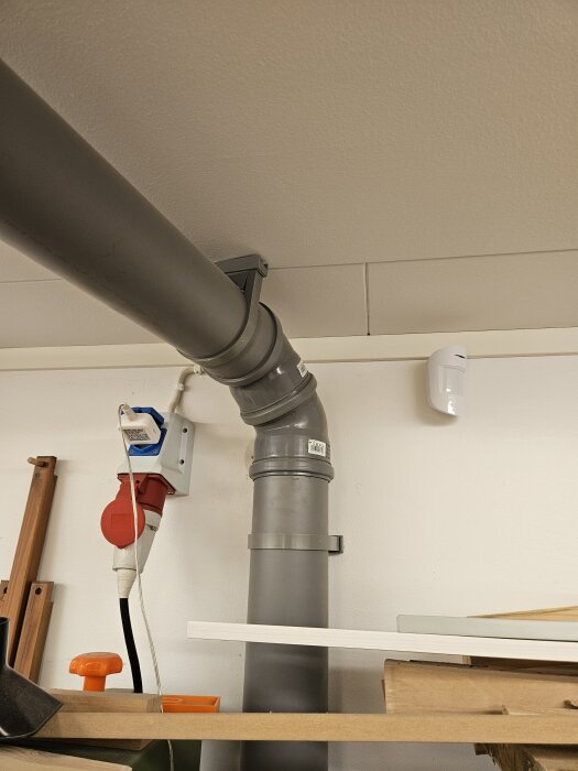Grått avloppsrör fäst vid väggen med klammer, elanslutning bredvid, flera verktyg och brädor runt omkring.