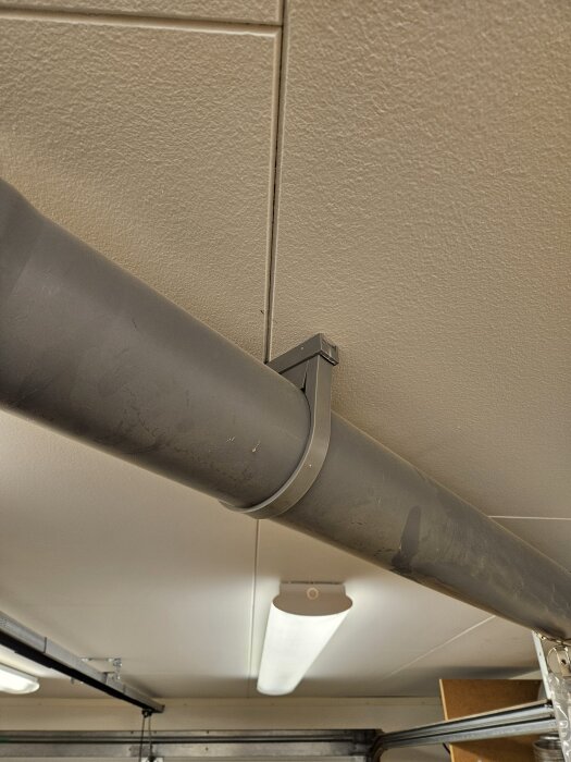 Grått avloppsrör fastsatt i taket med klammer och distanser, lysrör i bakgrunden.