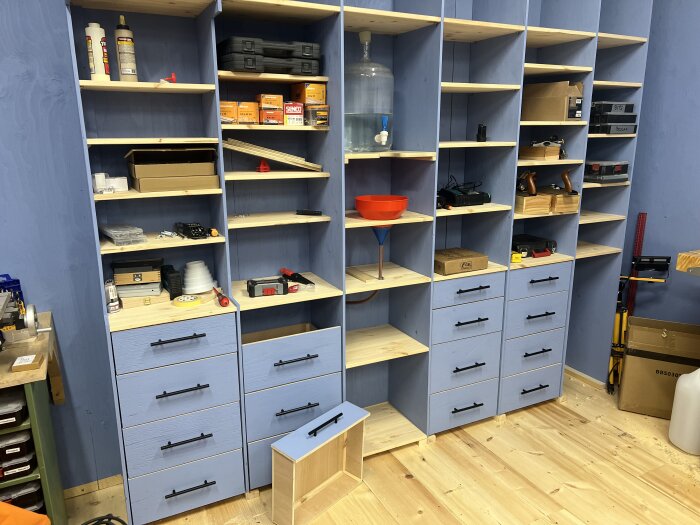 Blått förvaringssystem med hyllor och lådor fyllda med verktyg och byggmaterial i en verkstad.