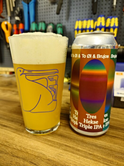 Ölglas med skummande öl bredvid en färgglad burk To Øl Tres Hekse Triple IPA på en arbetsbänk med verktyg i bakgrunden.