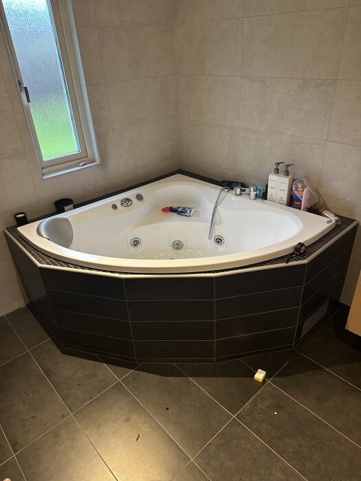 Inbyggt hörn-spabad i kaklat badrum, med svart plattor runt om och badrumsprodukter på kanten.