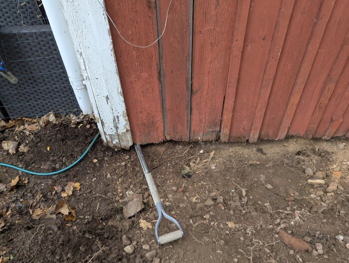 Spade under rödmålad trävägg med avskalad grund och grävd jord, där kanten på huset visar tecken på röta.