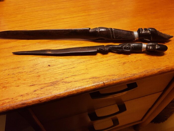 Två dekorerade brevknivar i trä, liggande på ett träbord. En är större, båda används för att öppna post.