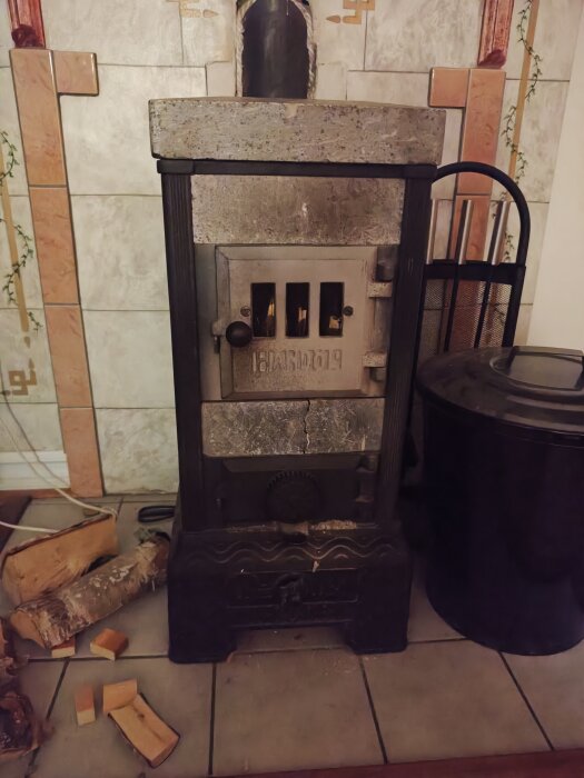 En äldre kamin av gjutjärn står på en kaklad yta med vedträn bredvid, omgiven av dekorativ kakelvägg.