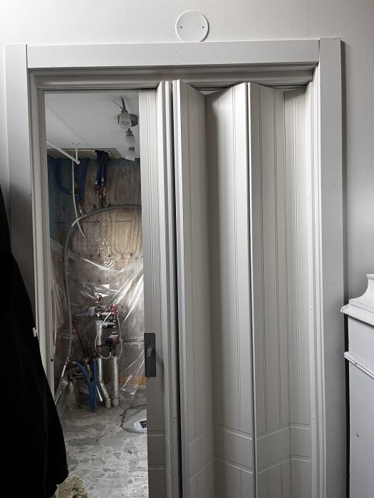 Vikdörr i dörröppning mellan hall och tvättstuga, med synlig eldosa ovanför dörren, och rör och plastskydd på väggen i bakgrunden.