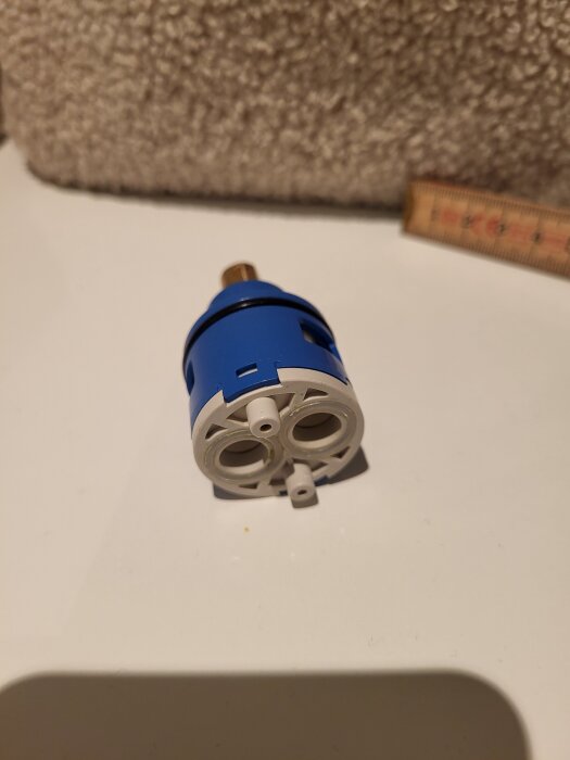 Blå och vit del av en vattenblandare med kopplingar synliga, placerad på en vit yta, med en linjal i bakgrunden för storleksreferens.