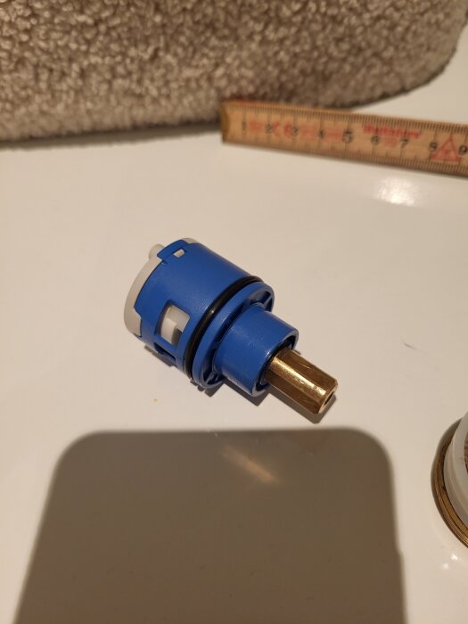 Blå och vit flödesventil för duschblandare på vit yta, med linjal i bakgrunden.