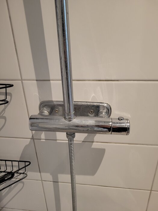 Kromad duschblandare monterad på vit kakelvägg, med en slang kopplad under blandaren.