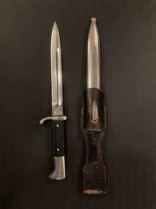 En dekorativ bajonett från mellankrigstiden bredvid sitt fodral. Hantaget är svart och metallen glänser, designad för prydnad snarare än funktion.