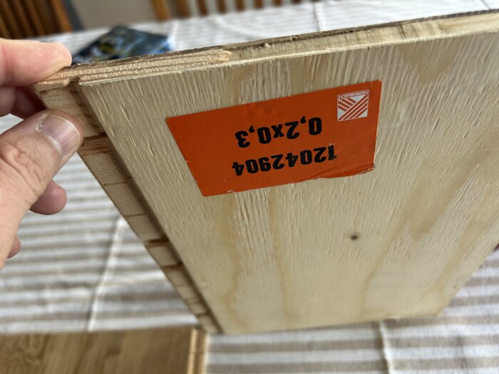 Närbild på en parkettgolvplanka med orange etikett från Hornbach, hålls upp av en hand över bordduk.