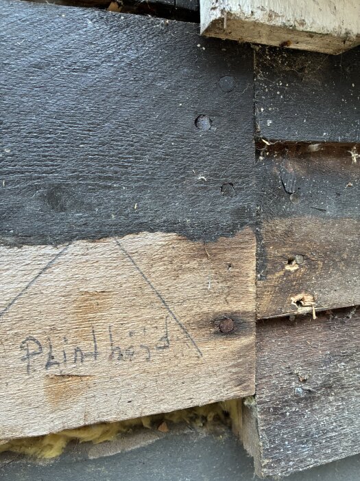 Närbild av träpanel med synliga skruvhuvuden och texten "plinthöjd" skriven på en träplanka. Fläckar och isolering syns också.