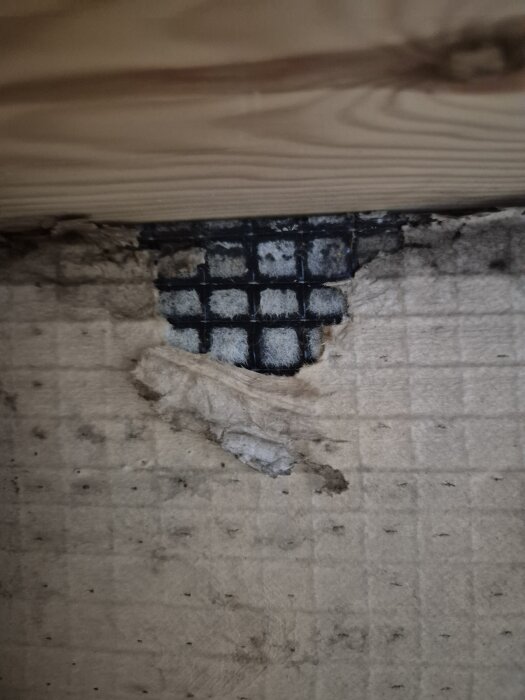 Skadad fasad visar blottad vindpapp och nätstruktur bakom tunn plastfilm, med trä ovanpå. Indikerar byggfel i konstruktionen.