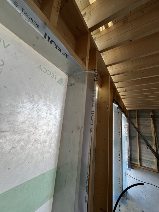 Bilden visar en trästomme i ett byggprojekt med plastfolie runt fönsteröppningar, där tejpad ångspärr syns tydligt.