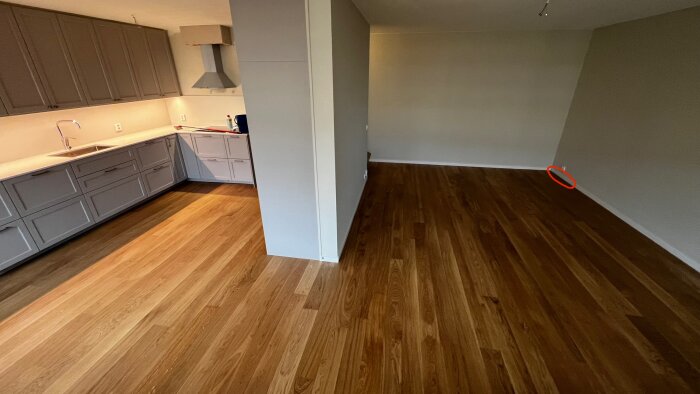 Vardagsrummet med trägolv visar en glipa markerad med en röd cirkel i hörnet nära köket, orsakad av sned golvläggning.