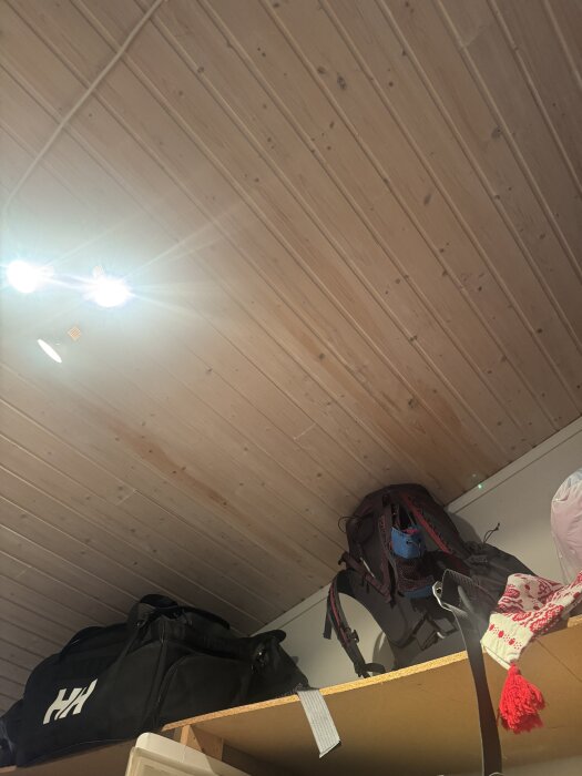 Trätak i förråd med mörka fläckar synliga; två resväskor och några kläder på en hylla under taket.