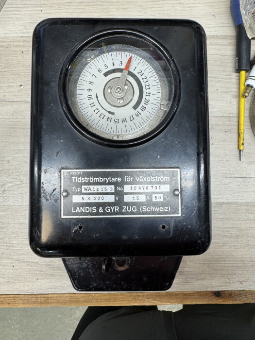 En svart tidströmbytare med ett urtavla och märkningen "Landis & Gyr Zug (Schweiz)" ligger på ett träbord.