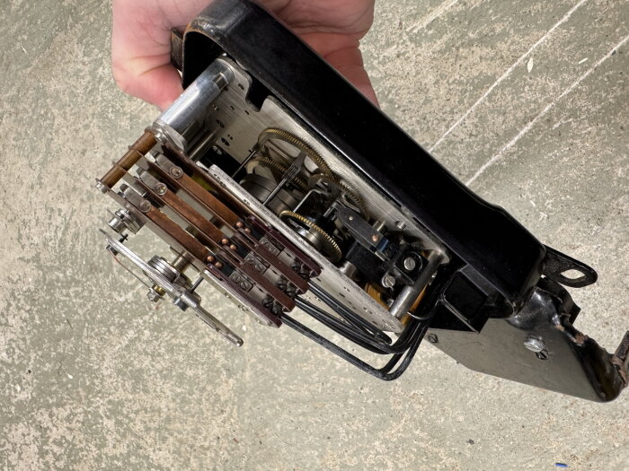 Närbild på insidan av ett mekaniskt tidur, med synliga kugghjul och kopparledningar, hålls i en hand över ett betonggolv.