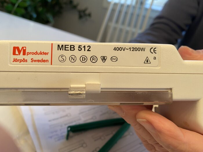 Termostat MEB 512 från LM produkter, 400V~1200W, hålls upp av en hand mot en suddig bakgrund med papper och green verktyg.