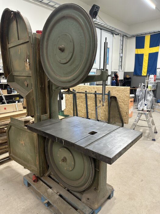Gammal bandsåg i en verkstadsmiljö, med ett svenskt flagga i bakgrunden, stående på en blå lastpall.