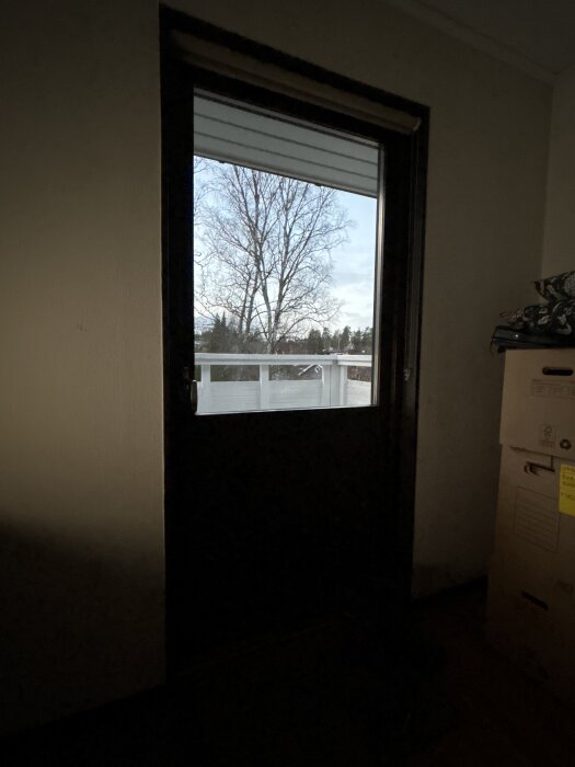 En brunbetsad dörr med glasruta och utsikt över ett träd och en altan. Till höger om dörren står flyttkartonger.