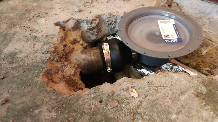 Golv uppbilat för installation av brunn ansluten till gjutjärnsrör, med fokus på muff och anslutning. Platta på mark utan våtrumscertifiering.