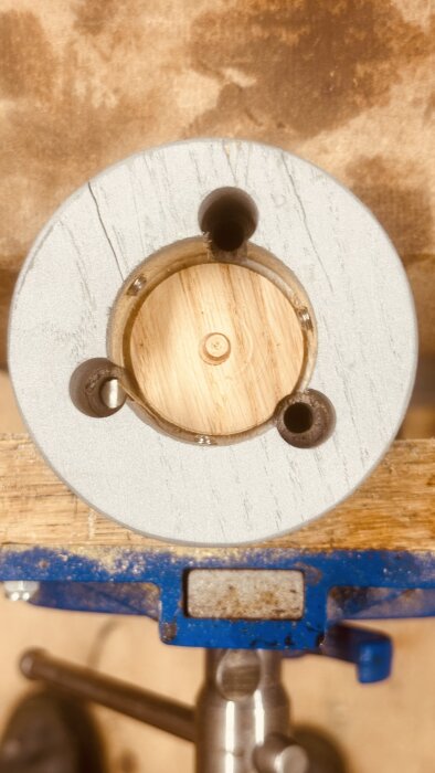 Metalladapter med tre låsskruvar och träplugg i mitten, monterad i skruvstäd på träbord.