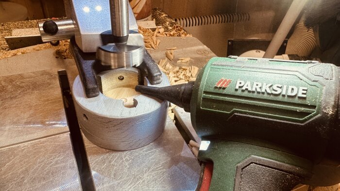 Borrmaskin borrar ett trästycke med hjälp av ett skruvstycke, omgiven av träspån. En Parkside-blåslampa syns till höger.