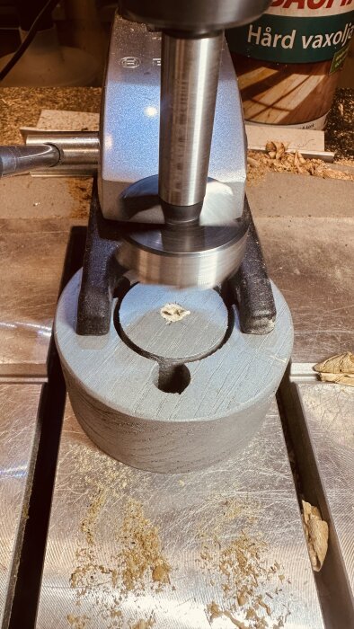 Borrmaskin borrar ett hål i en adapter med hjälp av ett Forstnerborr, omgiven av träspån. En burk med vaxolja syns i bakgrunden.