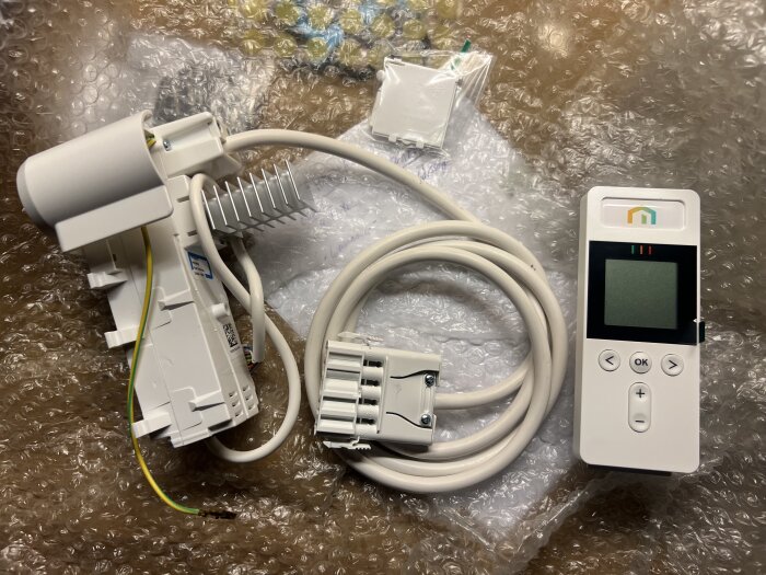 Termostatkit med elektronisk styrenhet, kablar och anslutningar för installation på radiator, placerat på bubbelplast.