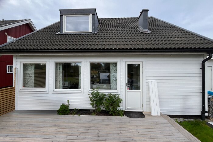Vitmålat hus med tre fönster och en dörr ut mot en träaltan. Svarta takpannor och växter vid entrén.