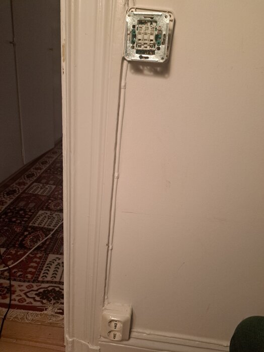 En vägg med ett oskyddat eluttag och vit kabeldragning, samt ett vägguttag nedanför.