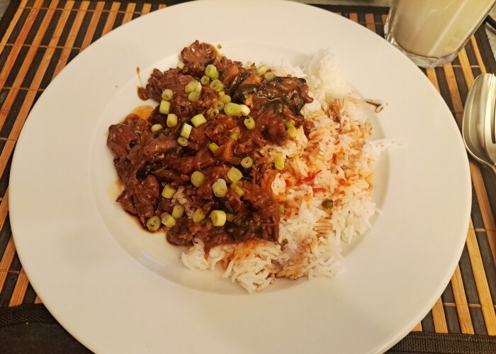 Tallrik med ris och köttgryta toppad med hackad purjolök, serverad på en bordstablett.