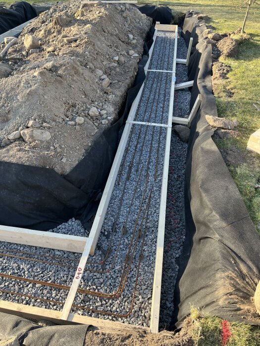 Grundläggning av växthus med grusfylld schakt, träram och armeringsjärn; omgiven av jordhögar och duken.