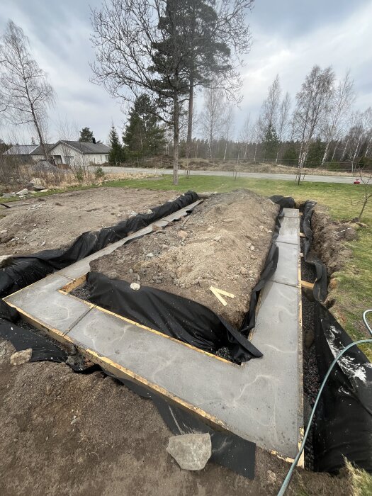 Grunden för ett växthus med nylagd betongsockel och lecablock syns på en gård i ett skogslandskap.