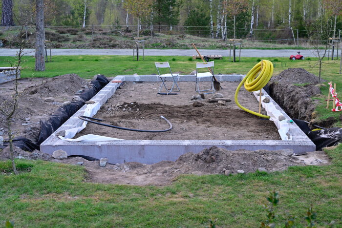 Grund för växthus med två stolar, verktyg och dräneringsrör på en gräsyta, omgiven av natur och träd.