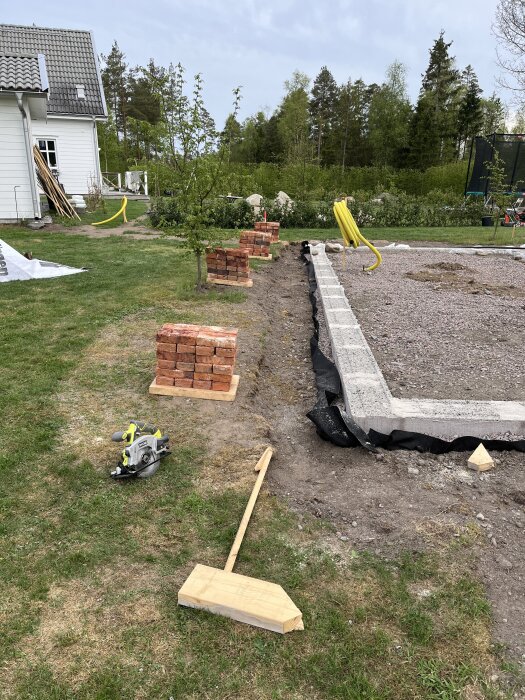Byggarbetsplats med staplade tegelstenar och en lecamur, omgiven av gräsmatta och träd, med ett hus i bakgrunden.