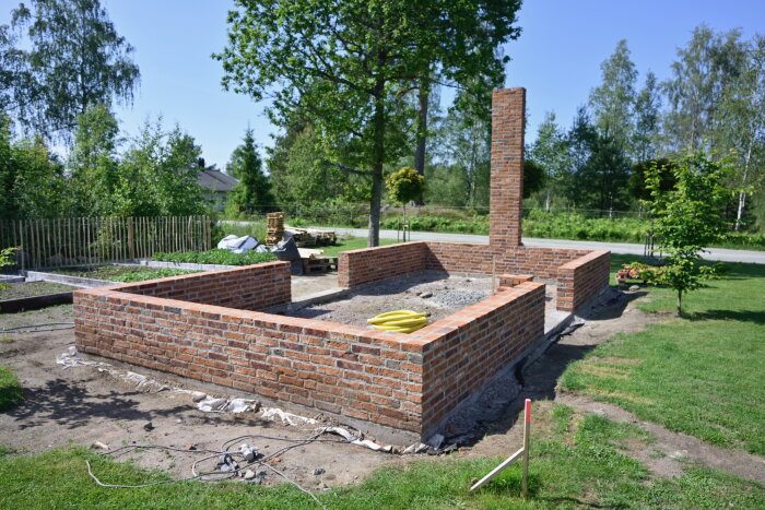 Murad lecamur kring grund för växthus under byggnation, omgiven av träd och gräsmatta, med en påse bananer ovanpå muren.