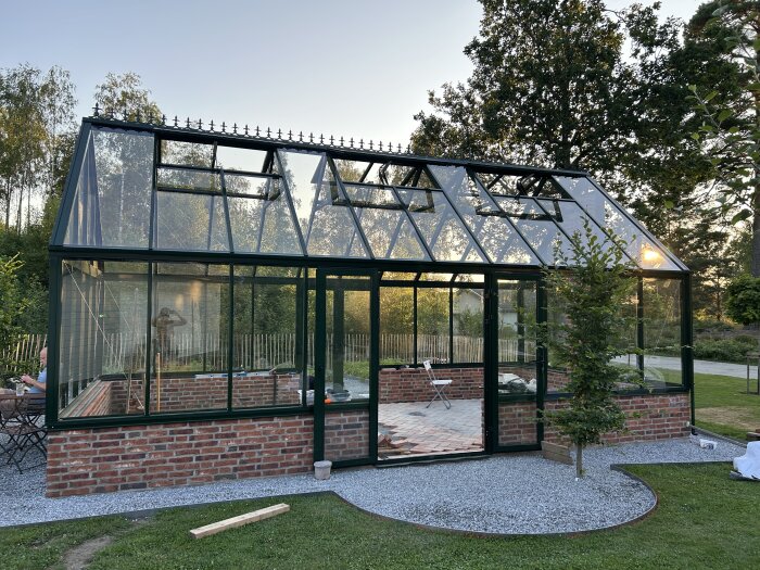 Växthus med tegelmur och glasväggar i trädgårdsmiljö, omgivet av grönska och grusgång.