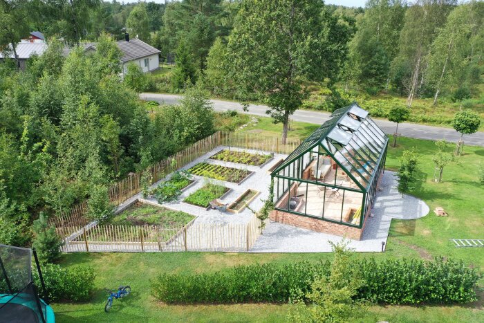 Färdigt växthus med grön ram bredvid odlingsbäddar i en trädgård. Omgiven av träd och en väg i bakgrunden.