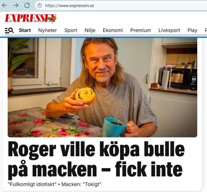En man med långt hår sitter vid ett bord och håller upp en bulle och en kopp kaffe i ett kök.