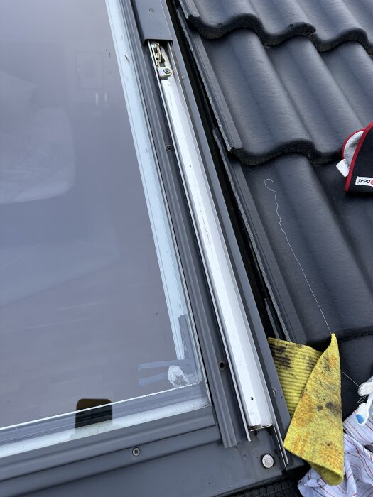Takfönster med skruvar och tätning synliga, samt takpannor bredvid; en smutsig trasa ligger på kanten av fönstret.