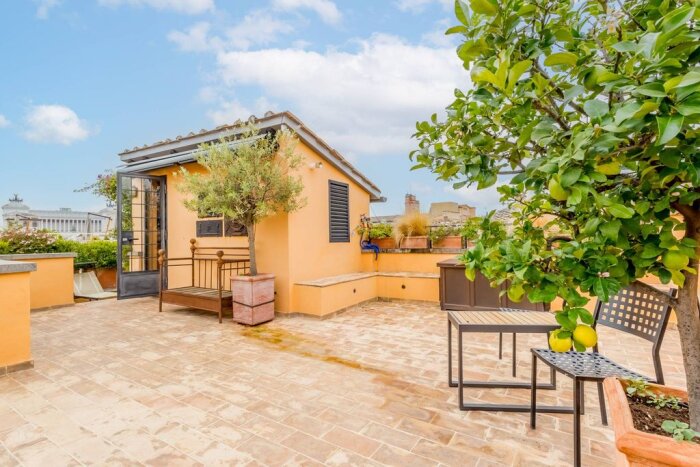 Takterrass med gul vägg, olivträd, citronträd, en bänk och bord med stolar. Vy över stadens byggnader i bakgrunden.