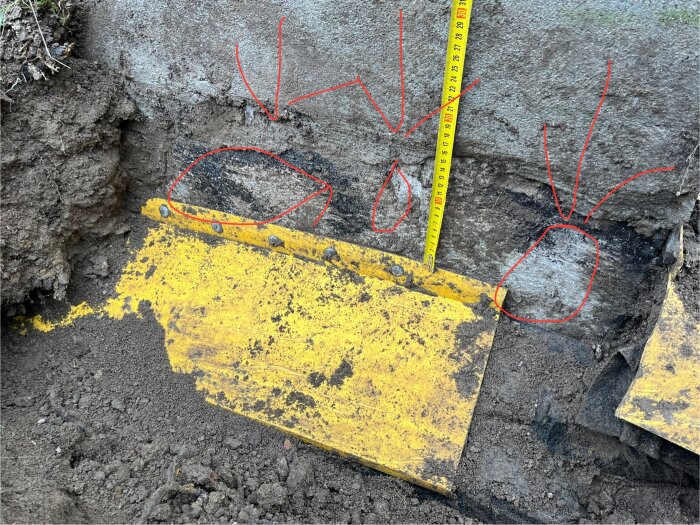 Gul täcklist vid grundmur med bristfällig installation, saknade delar, mätt med måttband. Marken visar tecken på dålig dränering och fuktskador.
