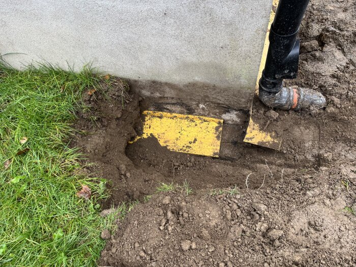 Grävd mark vid husgrund med synlig gul täcklist och rör, där dräneringsproblem undersöks och fuktskador vid marknivå noteras.