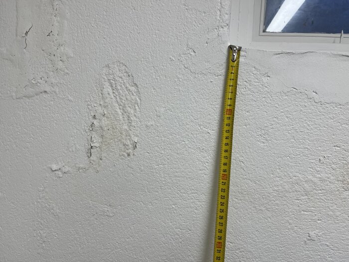 Bild av en vägg med fuktskador och ett måttband bredvid. Skadorna syns som avflagningar och missfärgningar på ytan.