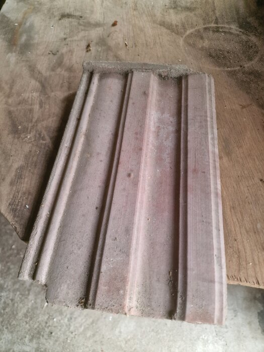 Gammal betongtakpanna liggande på ett träbord, delvis täckt av damm, med en klassisk enkelkupig profil.