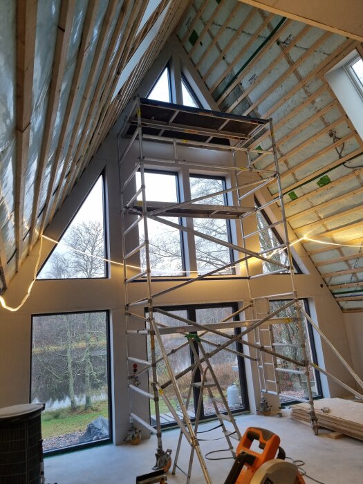 Byggnadsställning framför stora fönster och oisolerade takfack i en halvfärdig byggnad, med natur utanför.