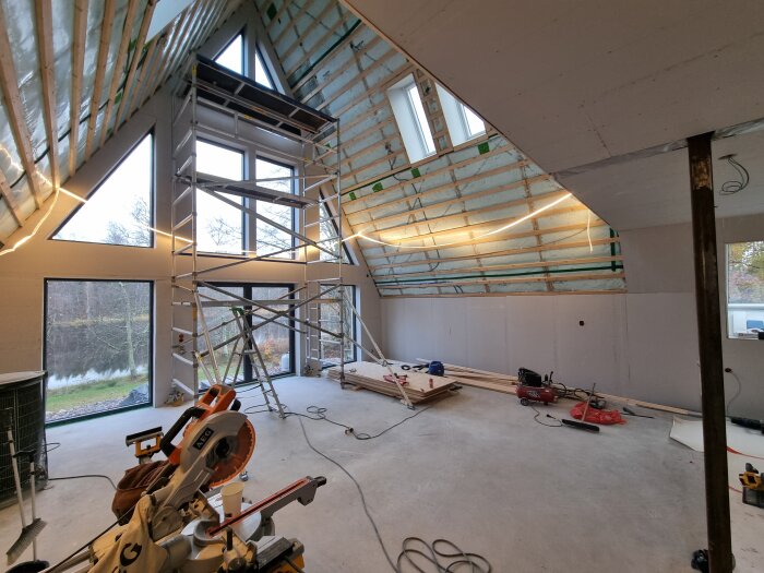 Interiör av ett hus under konstruktion med byggnadsställningar, stora fönster och verktyg spridda över golvet.