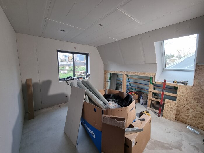 Renoveringsarbete i rum med gipsskivor på väggar och tak, verktyg och material utspridda, arbetsyta nära fönster; kartonger och byggmaterial synliga.