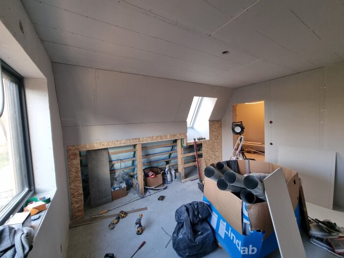 Renoverat rum med gipsskivor på väggar och tak, byggmaterial och verktyg på golvet, samt rör i låda och en bygglampa i hörnet.